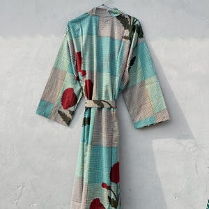May include: A long, patterned robe with a belt, hanging against a gray wall. The robe features a teal, brown, and white checkered pattern, accented with large red floral designs. The robe is hanging from a metal hanger.