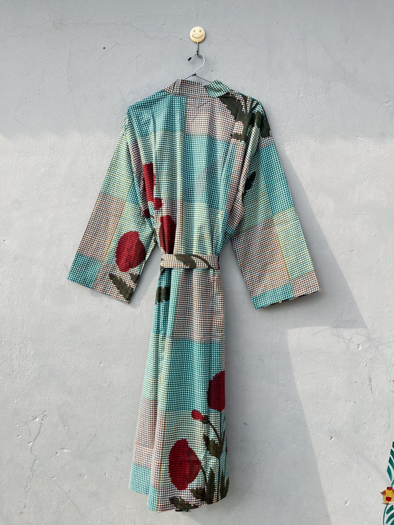May include: A long, patterned robe with a belt, hanging against a gray wall. The robe features a teal, brown, and white checkered pattern, accented with large red floral designs. The robe is hanging from a metal hanger.
