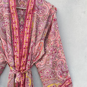 Puede incluir: Bata rosa con estampado de cachemira y cintur&oacute;n a juego. La bata presenta un dise&ntilde;o vibrante con tonos de rosa, morado y amarillo. La bata est&aacute; colgada en una percha de madera contra una pared gris.
