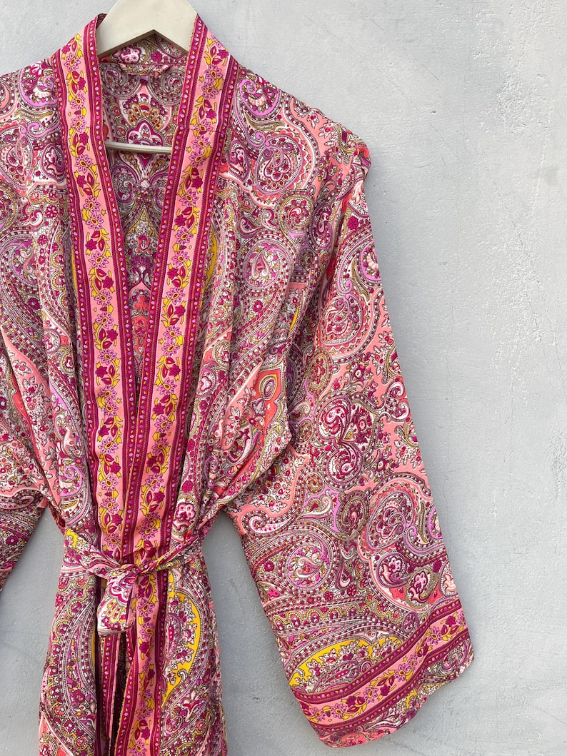 Puede incluir: Bata rosa con estampado de cachemira y cintur&oacute;n a juego. La bata presenta un dise&ntilde;o vibrante con tonos de rosa, morado y amarillo. La bata est&aacute; colgada en una percha de madera contra una pared gris.