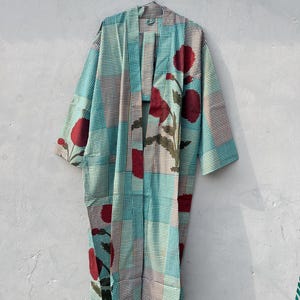 May include: A long, open-front robe with a patchwork design. The robe features a teal and white checkered pattern, accented with large red floral motifs. The robe is hanging on a metal hanger against a neutral wall.