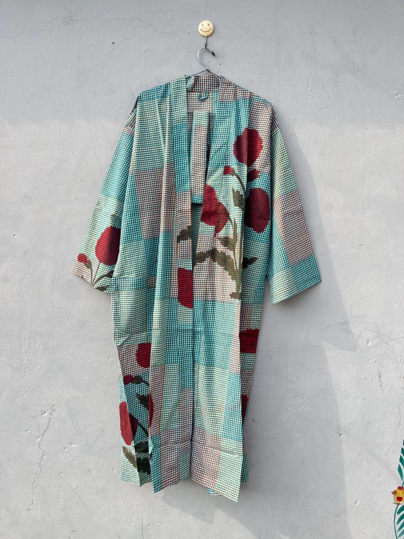 May include: A long, open-front robe with a patchwork design. The robe features a teal and white checkered pattern, accented with large red floral motifs. The robe is hanging on a metal hanger against a neutral wall.