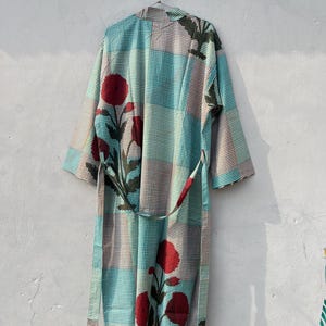 May include: A long, patterned robe with a belt, hanging against a light gray wall. The robe features a patchwork design of teal, brown, and white checks, with large red floral accents and dark green leaves. The belt is a matching pattern.