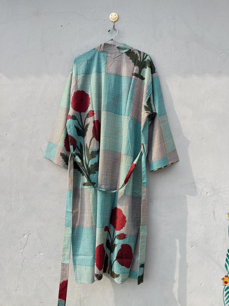 May include: A long, patterned robe with a belt, hanging against a light gray wall. The robe features a patchwork design of teal, brown, and white checks, with large red floral accents and dark green leaves. The belt is a matching pattern.