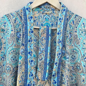 May include: A turquoise paisley patterned robe with blue and gold accents hangs on a wooden hanger. The robe features a detailed floral and swirl design. The robe's fabric is lightweight and appears to be silk or a similar material.