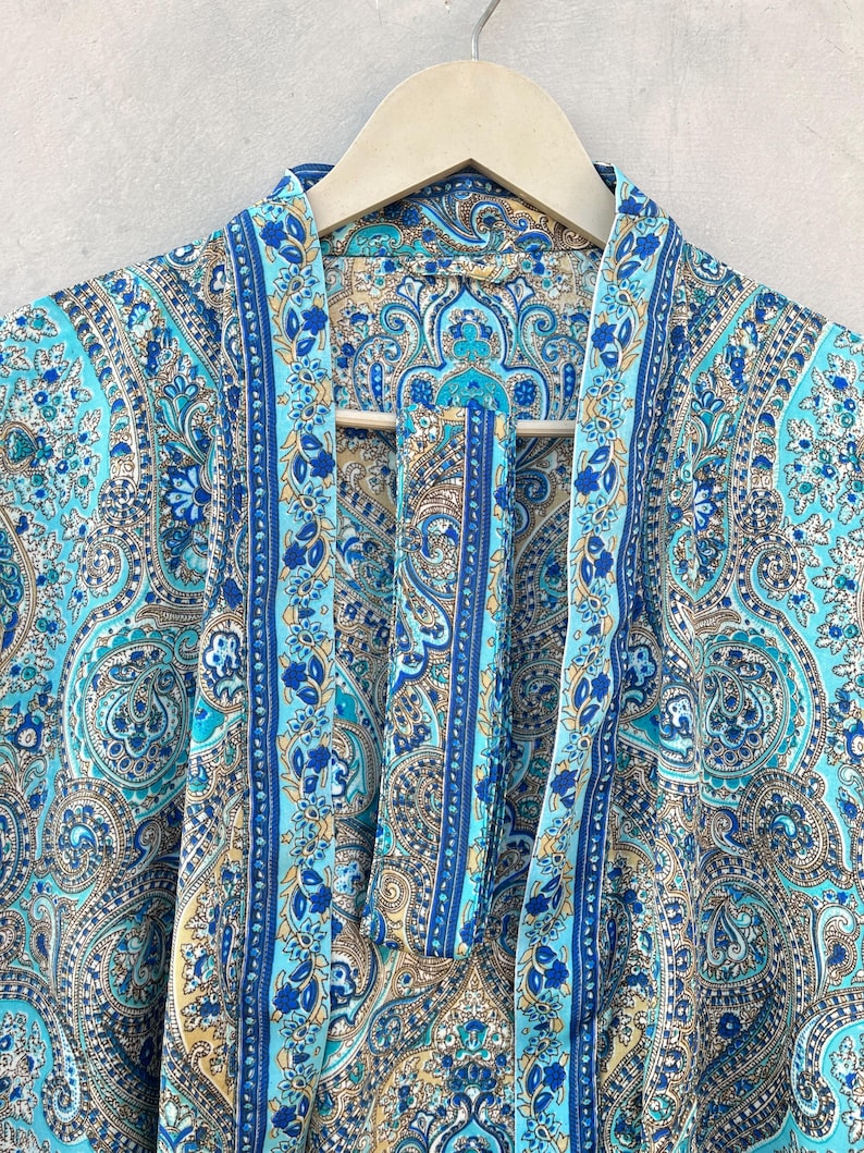 May include: A turquoise paisley patterned robe with blue and gold accents hangs on a wooden hanger. The robe features a detailed floral and swirl design. The robe's fabric is lightweight and appears to be silk or a similar material.