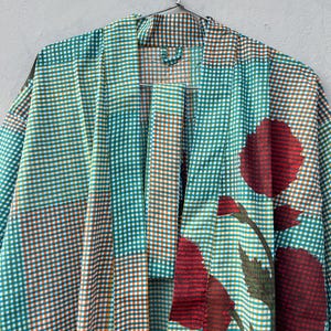 May include: A teal, orange, and white checkered kimono-style robe with large red floral accents. The robe is hanging on a silver metal hanger against a neutral background. The floral design includes red flowers and green leaves.