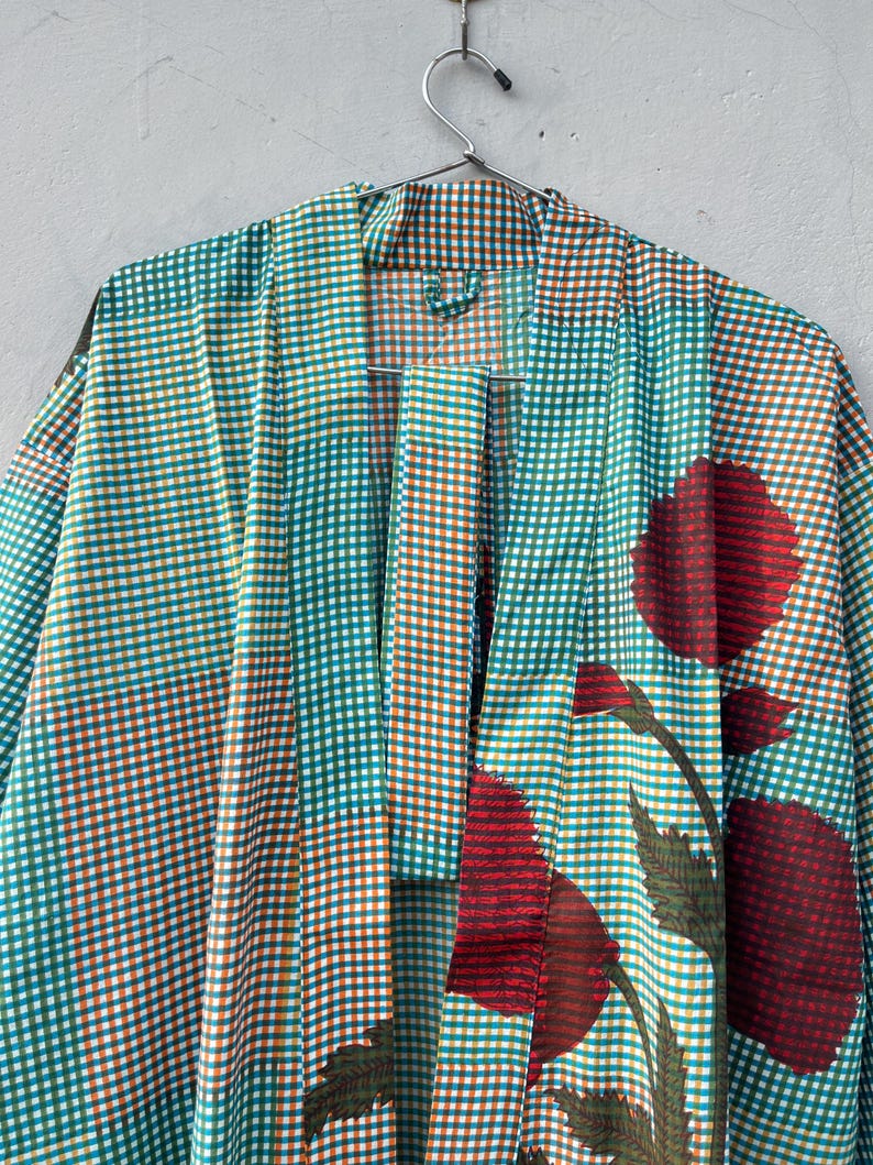 May include: A teal, orange, and white checkered kimono-style robe with large red floral accents. The robe is hanging on a silver metal hanger against a neutral background. The floral design includes red flowers and green leaves.