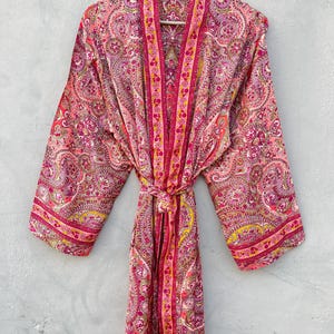Puede incluir: Bata rosa con estampado de cachemira y cintur&oacute;n a juego. La bata est&aacute; colgada de una percha de madera. Tiene un dise&ntilde;o floral en tonos rosa, amarillo y morado.
