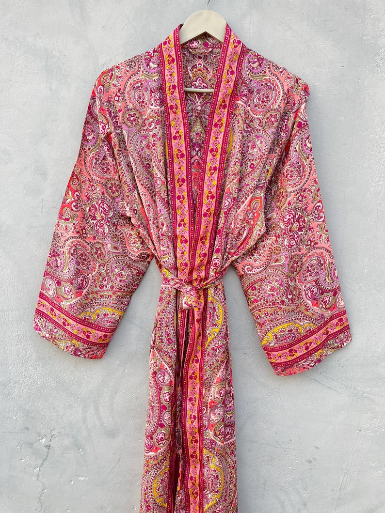Puede incluir: Bata rosa con estampado de cachemira y cintur&oacute;n a juego. La bata est&aacute; colgada de una percha de madera. Tiene un dise&ntilde;o floral en tonos rosa, amarillo y morado.