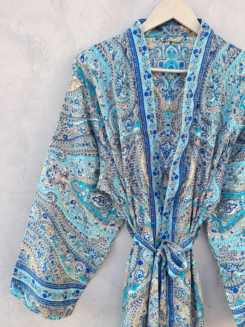 May include: A blue paisley patterned robe with a matching tie belt. The robe features shades of blue, teal, and tan. The robe is hanging on a wooden hanger against a neutral background.