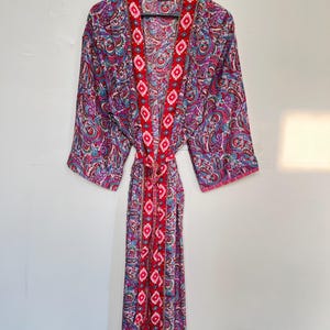 May include: A long-sleeved robe featuring a vibrant paisley pattern in shades of pink, blue, and purple. The garment includes a contrasting red front border and matching tie belt, both decorated with a repeating white and pink geometric diamond motif.