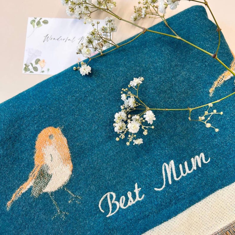 May include: Teal-colored throw blanket with an embroidered bird design and the words "Best Mum". The blanket is decorated with white flowers and a card that says "Wonderful Mum".