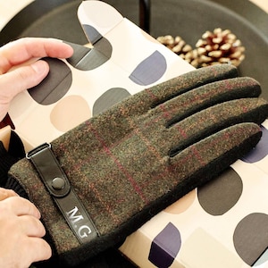 May include: A pair of olive green plaid gloves with black trim and a leather strap. The gloves have the initials "M.G." on the strap. The gloves are resting on a patterned gift box.