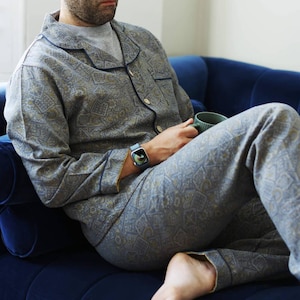 May include: A gray pajama set with a detailed pattern, featuring a button-down shirt and matching pants. The pajamas have navy blue trim and are worn with a gray t-shirt. A person is wearing a smartwatch and holding a green mug while sitting on a blue couch.