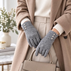 May include: A pair of grey knit gloves featuring three small decorative buttons with a plaid pattern along the outer wrist. The gloves have a textured, soft appearance and are worn with a beige coat and a structured beige handbag.