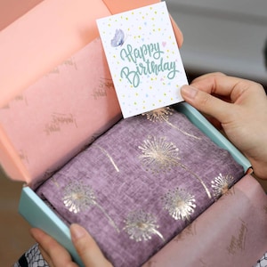May include: A gift box with a purple scarf featuring gold dandelion designs. The box contains a card that says "Happy Birthday" with a butterfly illustration. The box is light blue and pink.