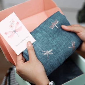 Könnte beinhalten: Ein teal-farbener Schal mit gestickten Libellenmustern wird in einer offenen Box präsentiert. Eine Karte mit einer Libellenillustration und dem Text "The joy is in the journey" wird neben dem Schal gehalten. Die Box hat ein rosa Innenleben.