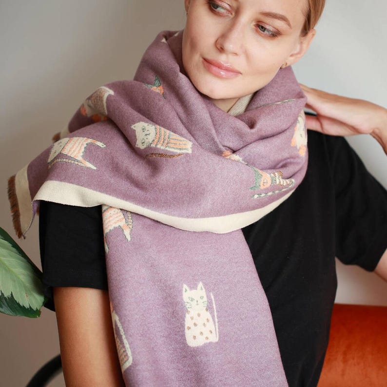 May include: A mauve scarf with a cream-colored border and cat illustrations. The scarf is wrapped around the neck and shoulders. The cat illustrations are in a whimsical style, with details in cream, orange, and green.