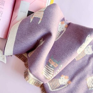 May include: A folded purple blanket with a repeating pattern of cartoon cats wearing glasses and striped shirts. The blanket has a cream-colored border and fringe along the edge. The word "ROJO" is visible on a tag.
