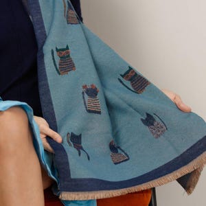 May include: A light blue scarf with a repeating pattern of cat illustrations. The cats are depicted in various poses and are wearing glasses. The scarf has a dark blue border and fringe.
