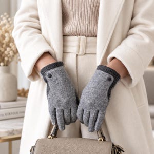 Könnte beinhalten: Ein Paar hellgraue Handschuhe in Wolloptik mit einem dunkelgrauen Fischgrätenmuster am Bündchen und einem kleinen runden Knopfdetail am äußeren Handgelenk. Die Handschuhe sind in einer klassischen Fünf-Finger-Form gestaltet.
