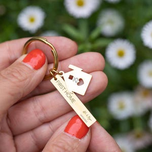 Könnte beinhalten: Ein goldfarbener Schlüsselanhänger mit einem hausförmigen Anhänger und einem rechteckigen Anhänger. Der Anhänger trägt die Aufschrift "FIRST HOME" und enthält ein Datum und Namen. Der Hausanhänger hat einen Herz-Ausschnitt und die Zahl "25".