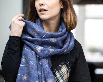 May include: A blue scarf with a dandelion pattern. The scarf is made of a lightweight fabric and features a repeating design of gold-colored dandelions and white stems. The scarf is wrapped around the neck.