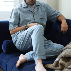 May include: Light blue pajama set with a grid pattern. The set includes a button-down shirt and matching pants. The person is wearing a watch and holding a green mug. The person is sitting on a dark blue couch.