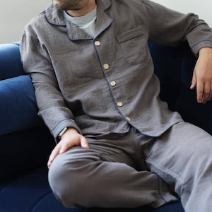 May include: Gray pajama set with navy blue trim and buttons. The long-sleeved top has a chest pocket. The person is sitting on a blue couch. The pajama set is made of a soft, comfortable material.