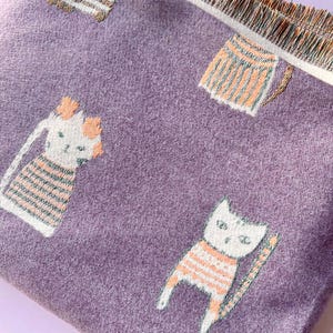 May include: A close-up of a purple fabric with a repeating pattern of cartoon cats wearing orange and white striped shirts. The fabric has a fringed edge.