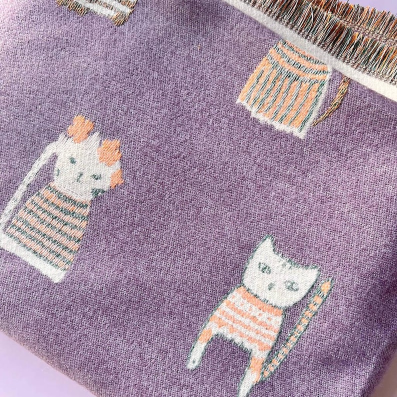 May include: A close-up of a purple fabric with a repeating pattern of cartoon cats wearing orange and white striped shirts. The fabric has a fringed edge.