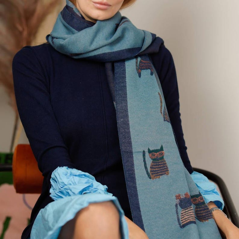 May include: A blue scarf with a cat pattern. The scarf has a teal background with dark blue borders and cat illustrations in brown and tan. The person is wearing a navy blue dress and a light blue blouse.