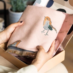 May include: A pink throw blanket featuring a robin bird design, presented in a cardboard box. The blanket has a fringed edge and a grey border. The robin is illustrated in shades of orange, brown, and grey.