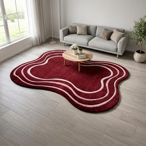 Könnte beinhalten: Ein burgunderfarbener Teppich mit einer einzigartigen, organischen Form. Der Teppich hat ein wellenförmiges Design mit hellrosa Streifen. Der Teppich befindet sich in einem Wohnzimmer mit einem grauen Sofa, einem Holztisch und einer Topfpflanze.