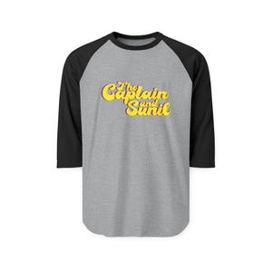 May include: A gray raglan baseball tee with black sleeves. The shirt features the text "The Captain and Sunil" in yellow, retro-style lettering. The shirt is a casual style.