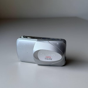 May include: Silver Olympus Camedia D-560 Zoom digital camera. The camera has a rounded design with a lens on the front and a small screen on the back. The Olympus logo and camera model are visible on the front.