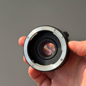 Op de afbeelding: Een zwarte en zilveren camera lens adapter met een heldere glazen lens. De adapter heeft een cirkelvormig ontwerp met verschillende schroeven en een kleine hendel. De adapter wordt in een hand gehouden tegen een grijze achtergrond.
