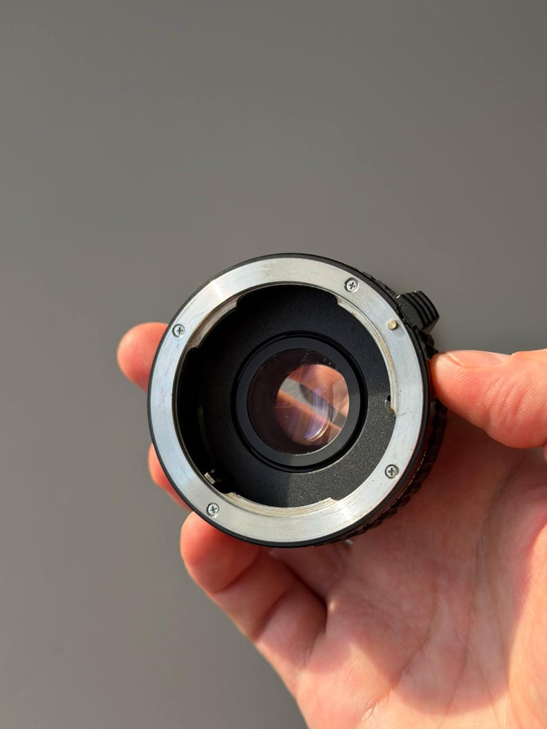 Op de afbeelding: Een zwarte en zilveren camera lens adapter met een heldere glazen lens. De adapter heeft een cirkelvormig ontwerp met verschillende schroeven en een kleine hendel. De adapter wordt in een hand gehouden tegen een grijze achtergrond.