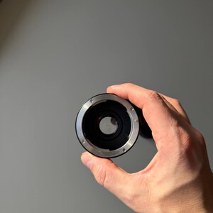 Op de afbeelding: Een zwarte en zilveren camera lens adapter. De adapter wordt in een hand gehouden en heeft een cirkelvormig ontwerp met een centrale opening en kleine schroeven. De achtergrond is een effen grijze muur.