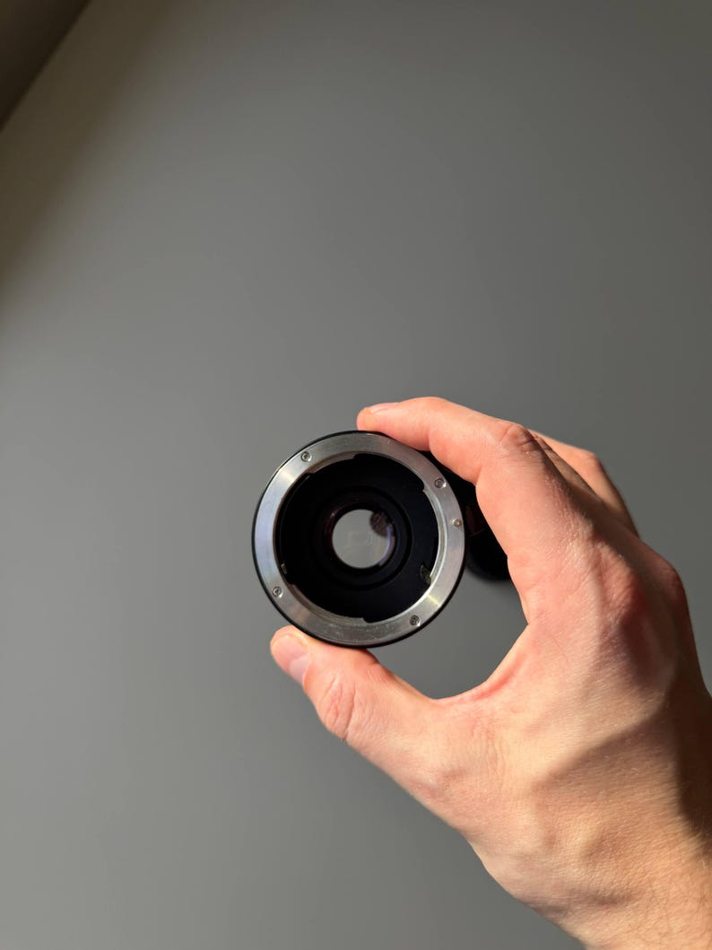Op de afbeelding: Een zwarte en zilveren camera lens adapter. De adapter wordt in een hand gehouden en heeft een cirkelvormig ontwerp met een centrale opening en kleine schroeven. De achtergrond is een effen grijze muur.