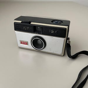 May include: Vintage Kodak Instamatic 124 camera. The camera has a black top and a silver body with the Kodak logo and the number 124. A black strap is attached to the side. The camera is a classic piece of photography equipment.