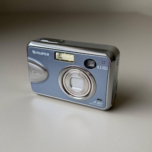 May include: A silver and light blue Fujifilm A360 digital camera featuring a Fujinon zoom lens, a built-in flash, and a 4.1 megapixels label. The compact device has a metallic finish with clearly visible branding and model text on the front panel.