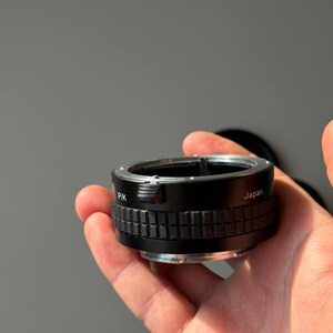 Op de afbeelding: Een zwarte en zilveren camera lens adapter met de tekst "PK" en "Japan". De adapter heeft een geribbeld, getextureerd ontwerp rond het midden. Het wordt in een hand gehouden tegen een grijze achtergrond. Voor fotografie.