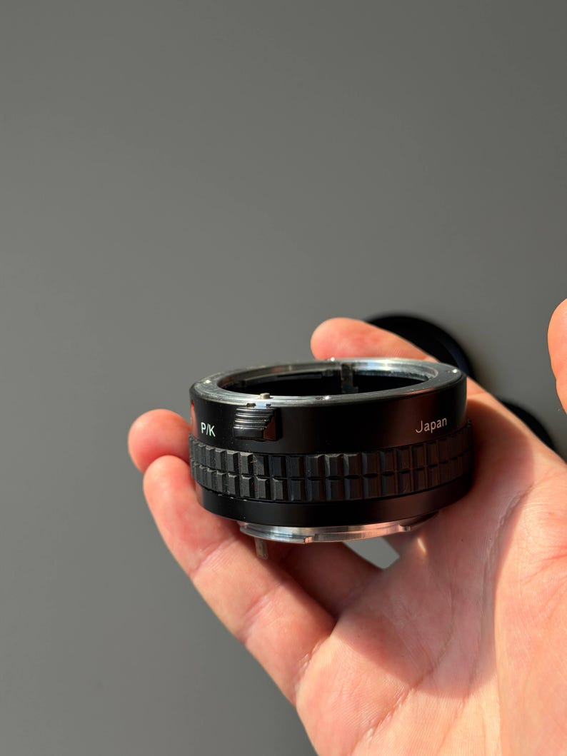 Op de afbeelding: Een zwarte en zilveren camera lens adapter met de tekst "PK" en "Japan". De adapter heeft een geribbeld, getextureerd ontwerp rond het midden. Het wordt in een hand gehouden tegen een grijze achtergrond. Voor fotografie.