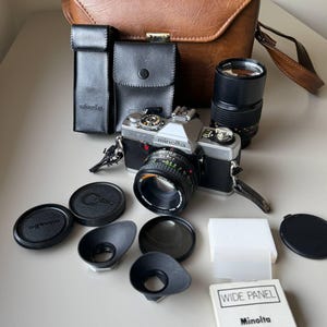 Puede incluir: Un kit de cámara Minolta vintage con una cámara plateada y negra con lente, varios accesorios y una funda de transporte de cuero marrón. Incluye tapas de lente, visores y una caja "WIDE PANEL".