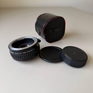Op de afbeelding: Een zwarte teleconverter met de tekst "Auto 2x Tele-Converter" en twee zwarte lensdoppen. Er is ook een zwarte leren etui met rode stiksels aanwezig. De items liggen op een wit oppervlak.