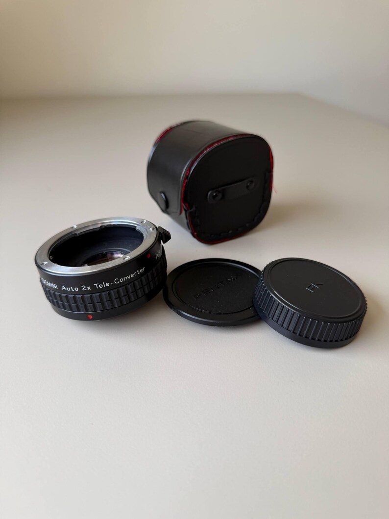 Op de afbeelding: Een zwarte teleconverter met de tekst "Auto 2x Tele-Converter" en twee zwarte lensdoppen. Er is ook een zwarte leren etui met rode stiksels aanwezig. De items liggen op een wit oppervlak.