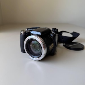 Peut inclure: Appareil photo numérique Olympus noir avec un anneau d'objectif argenté et une sangle noire. Le nom de la marque "Olympus" est imprimé sur le dessus. Le bouchon d'objectif est également visible.