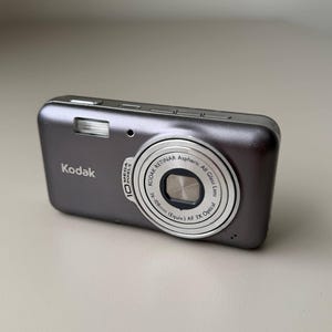 May include: A charcoal gray Kodak digital camera featuring a silver lens barrel with text reading Kodak Retinar Aspheric All Glass Lens 36-108mm Equiv AF 3X Optical and 10 Mega Pixels. The device has a rectangular body with a flash unit on the front.